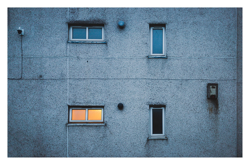 A picture of an apartment block with a wall and windows The block is neglected and empty looking, but one of the windows is lit