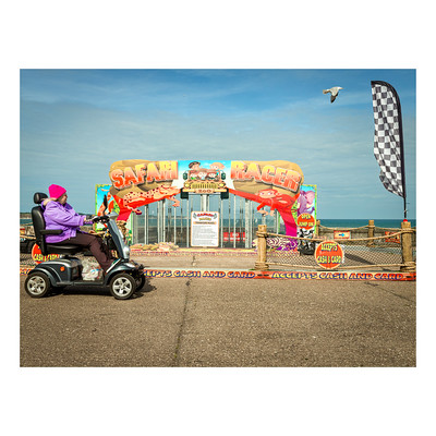 A side on view of a woman dressed elegantly in purple with a matching hat driving her mobility scooter. The scooter is being driven on the promenade, with the horizon between sea and sky visible. BEhind the woman is a childrens fairground ride called Safari Racer with pictures of cartoon lions and other jungle animals. At the top right of the image a seagull is flying, the same direction as the woman is driving as if they were racing each other.